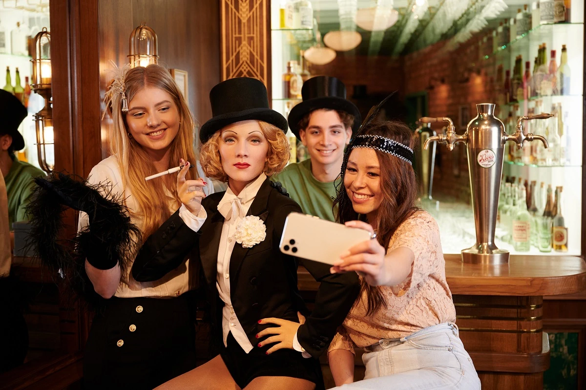 Madame Tussauds Berlin, Wachsfiguren, Marlene Dietrich sitzt im schwarzen Tanzanzug, schwarzem Hut und Zigarette in der Hand an einer Bar. Drei Jugendliche machen ein Selfie mit ihr.
