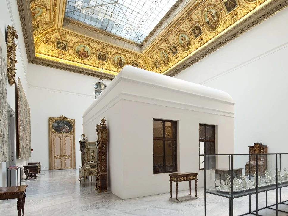 Blick in die Schausammlungen im MAK Museum Wien. Ein weißes Haus steht in einer großen Halle. Die Decke ist mit einem goldenen Rahmen und Fenstern versehen. Verscheidene Ausstellungsstücke stehen im Raum verteilt.