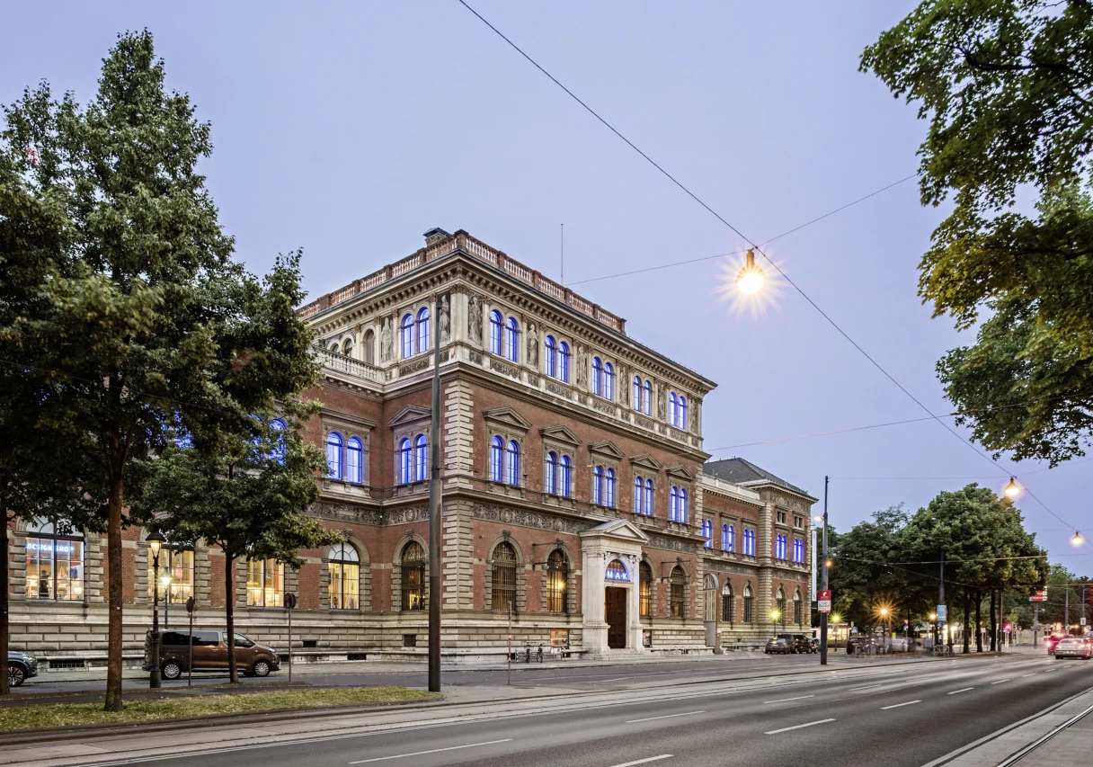 Außenansicht des MAK Museum Wien zur blauen Stunde. Der Ring als große Straße ist leer und die Laternen leuchten schon in der Höhe. In den Fenstern des Museums brennt Licht.