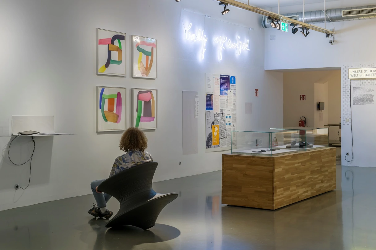 Blick in das MAK Design Lab. Eine Frau sitzt auf einem Stuhl und schaut auf vier quadratische bunte Bilder, die an der Wand hängen.