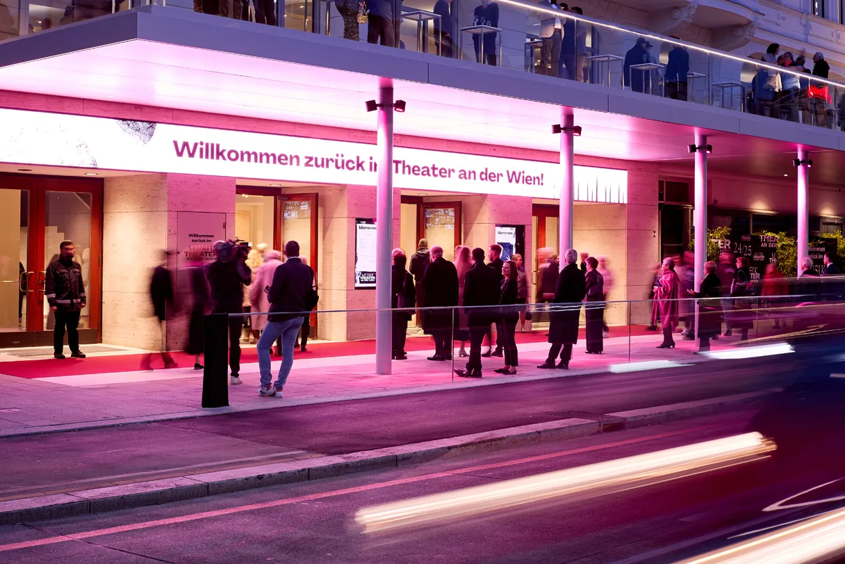 Exterior view of the Theater an der Wien. “Welcome back to the Theater an der Wien” is written at the entrance. People stand in front of the entrance or walk past.