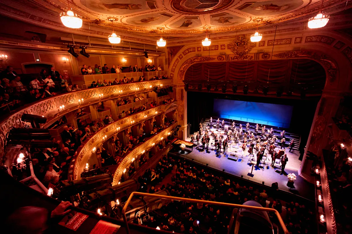 View into the theater hall from the upper tier. You can see the stage with artists and the audience sitting in the rows.