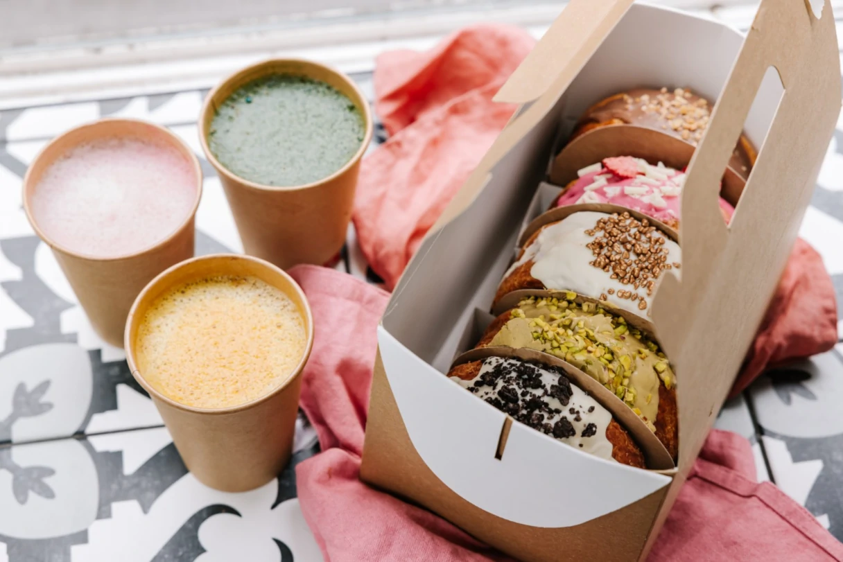 New York Rolls, Vienna, a paper box with five half New York Rolls stands on the table. Next to it are three cups filled with hot drinks