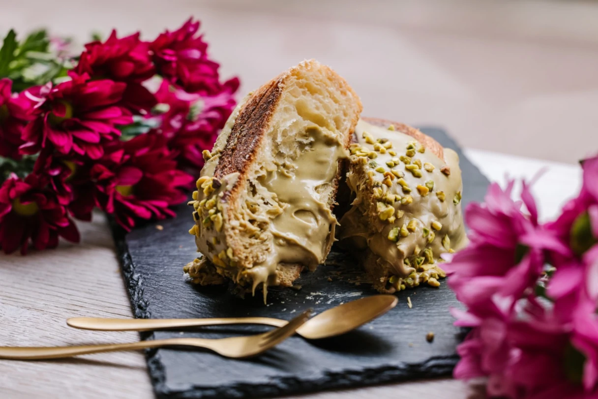 New York Rolls, Wien, ein Pistazien Roll liegt aufgeschnitten auf einer schwarzen Schieferplatte. Davor liegt goldenes Besteck und rechts und links pinke Blumen