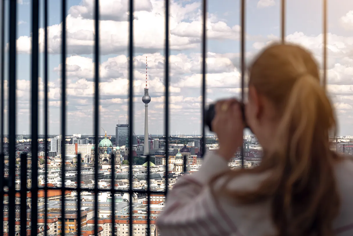 Frau mit Pferdeschwanz ist von hinten am rechten Bildrand zu sehen. Sie hat eine Kamera in der Hand und schaut durch die Gitter auf der Aussichtsplattform des Panoramapunkts. Man sieht den Fernsehturm zwischen den Streben und andere Berliner Sehenswürdigkeiten.