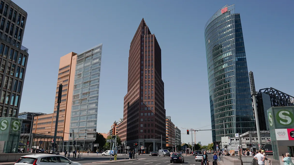 Blick auf den Panoramapunkt von der Leipziger Straße aus. Rechts steht das DB Gebäude, links ein anderes spitzes Gebäude. Sonniger Tag mit blauem Himmel.