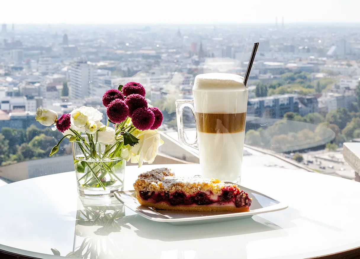 Ein Tisch im Café mit einem Kuchenteller, Kaffee und Blumen. Im Hintergrund kann man Berlin von oben erkennen.