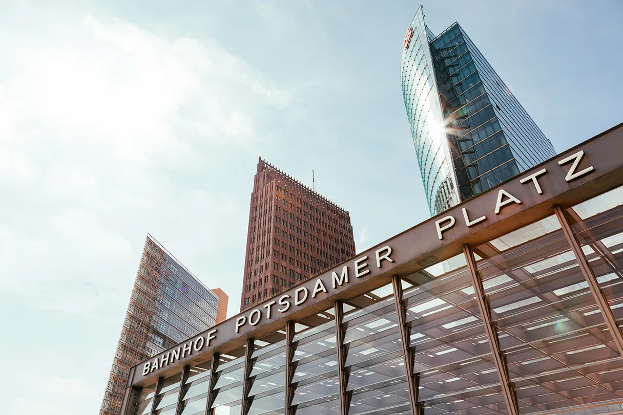 Dachkonstruktion des S-Bahnhof Potsdamer Platz. Im Hintergrund sind der Panoramapunkt und zwei andere Hochhäuser zu sehen. Die Sonne spiegelt sich in einem Hochhaus.