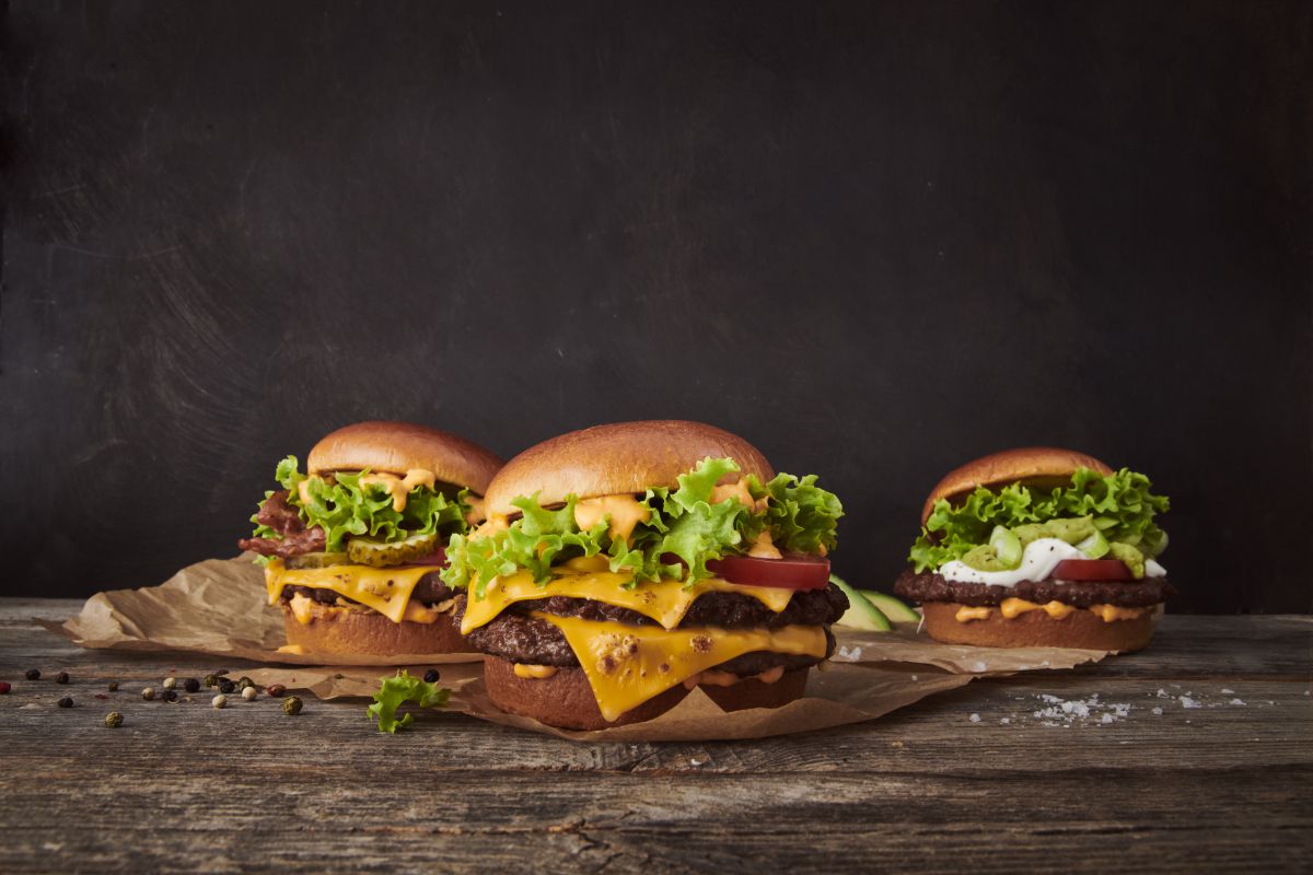 drei Burger liegen auf Papier und einem dunklen Holztisch. Viel Käse, grüne Salatblätter und Fleisch.