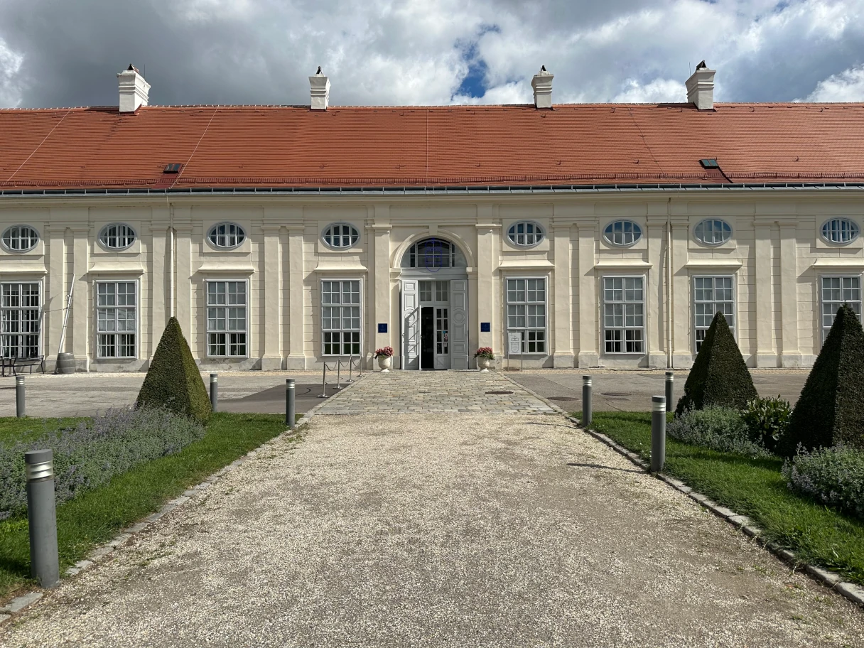 View of the entrance area of the Augarten Porcelain Museum in Vienna. A gravel path is lined to the right and left by lawns and lacy green fir trees.