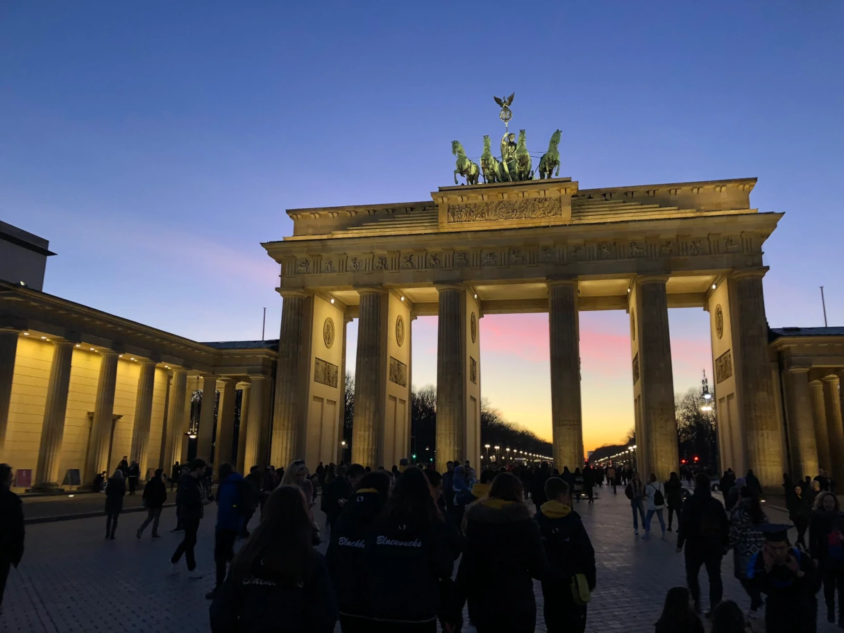 Brandenburg Gate at sunset, many people as black silhouettes in front of it, the sky is colored in blue, pink and yellow