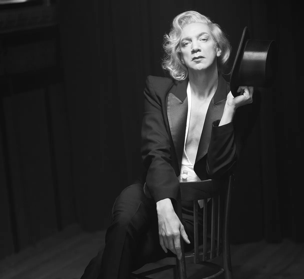 Renaissance Theater Berlin, Schauspieler als Marlene Dietrich sitzt im schwarzen Anzug auf einem Stuhl und hält einen Hut in der Hand, schwarz weißes Bild