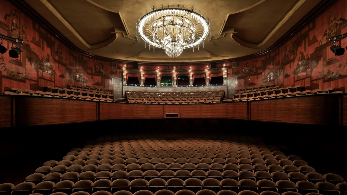 Blick in das Renaissance Theater Berlin. Riesiger Kronleuchter an der Decke, man sieht die Intarsien an den Wänden, rechts und links der Logenplätze. Im Parkett sieht man die vielen Sitzplätze. Farblich in rot, braunen Tönen gehalten.