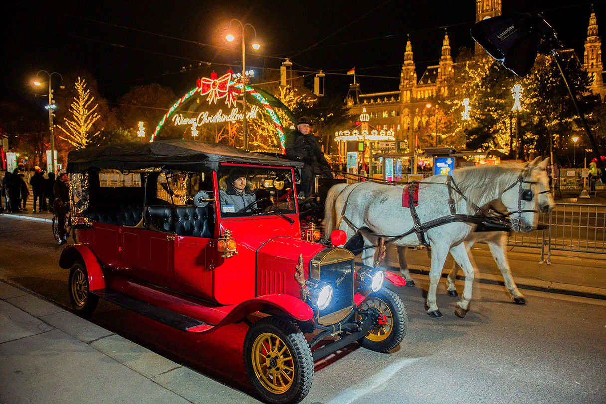 Oldtimer Auto von Ring Tours Vienna steht auf der Straße vorm Burgtheater und Rathausplatz Wien. Ein Fiaker mit weißen Schimmeln fährt neben dem Oldtimer.