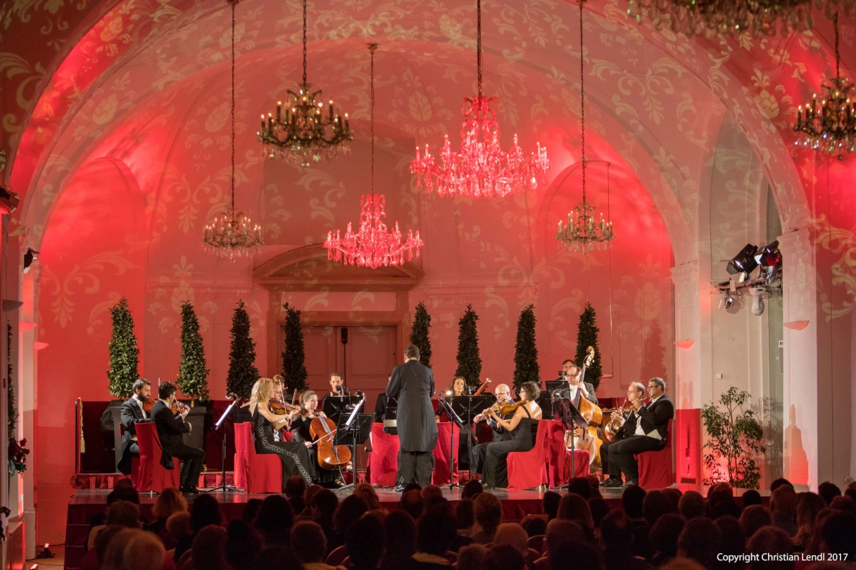 Schloss Schönbrunn Konzerte, Bühne in der Orangerie im Schloss Schönnbrunn, rot angeleuchtet, Kronleuchter hängen von der Decke, Blick vom Saal auf das Orchester