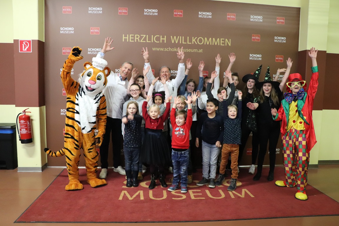 SchokoMuseum Vienna, group photo, children and adults standing at the entrance to the museum in front of the photo wall, mascot standing to the left of the picture, everyone laughing and raising their arms in the air