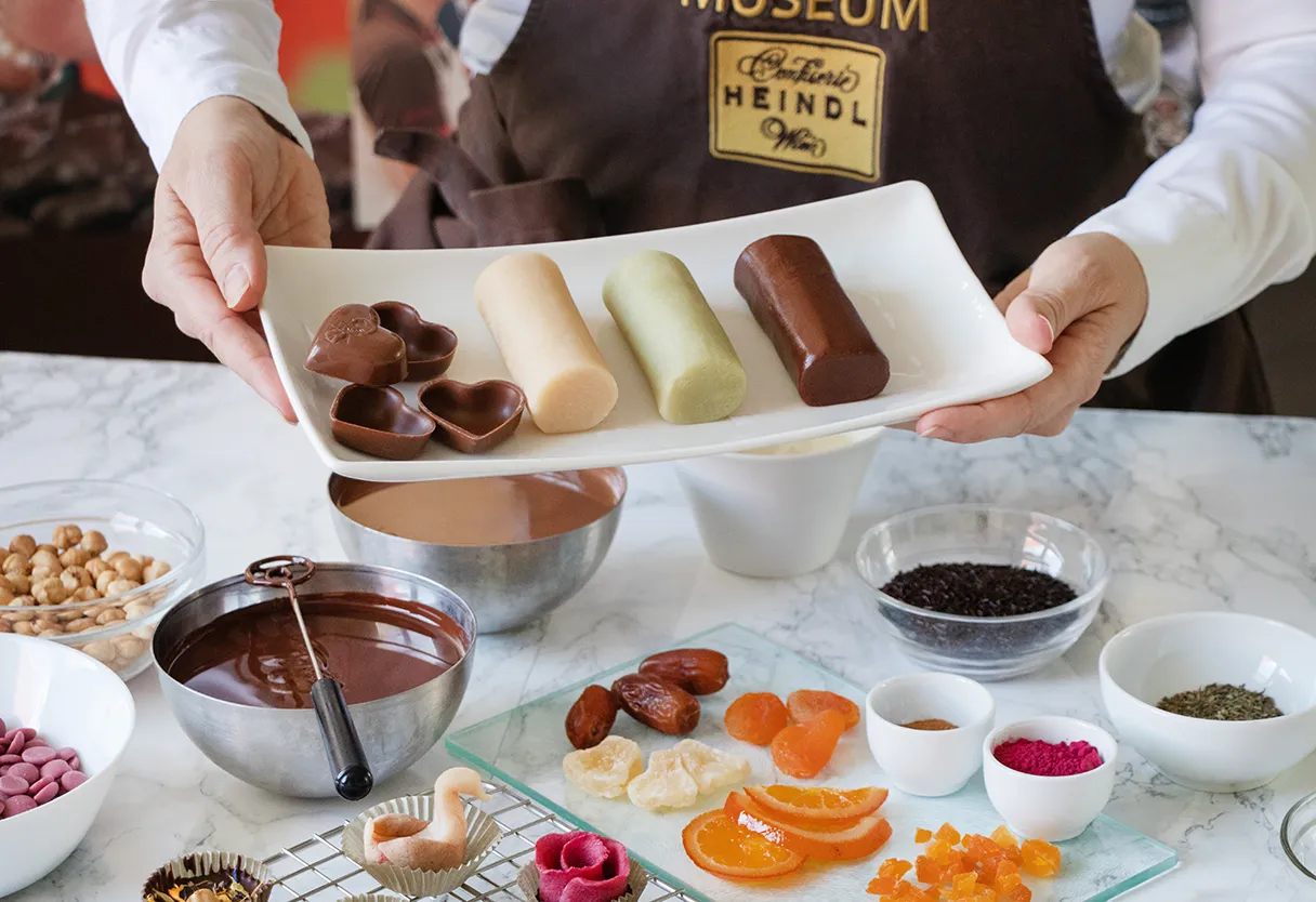 Pralinenworkshop im SchokoMuseum Wien. Auf einem weißen Tablet liegen selbstgemachte Pralienen, die von einem Mitarbeiter über einen Tisch gehalten wird. Auf dem Tisch ist verschiedenes Obst, Schokolade und andere Zutaten für den Pralinenworkshop ausgestellt.