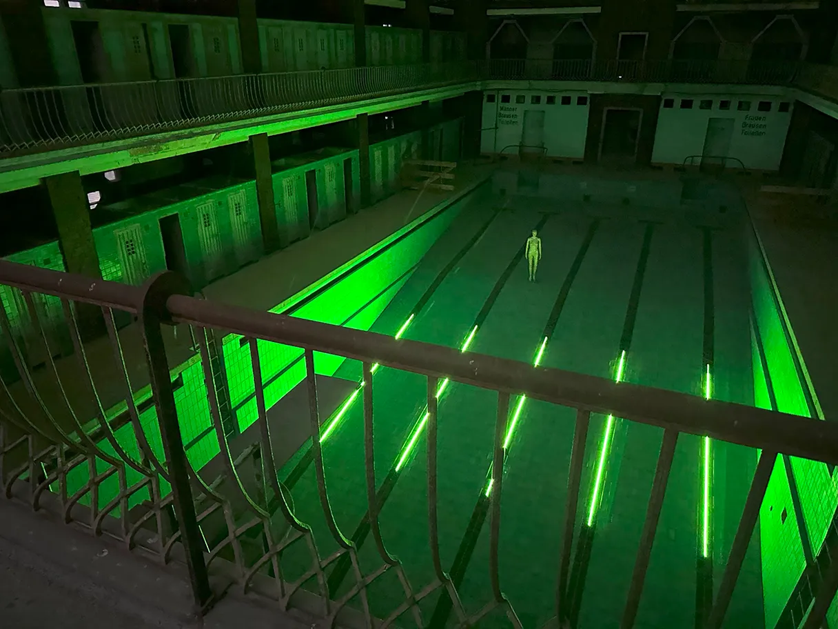 Stadtbad RELOADED: AMBILIGHT, grün beleuchtetes leeres Schwimmbad, Blick von oben, eine Person steht im Schwimmbecken