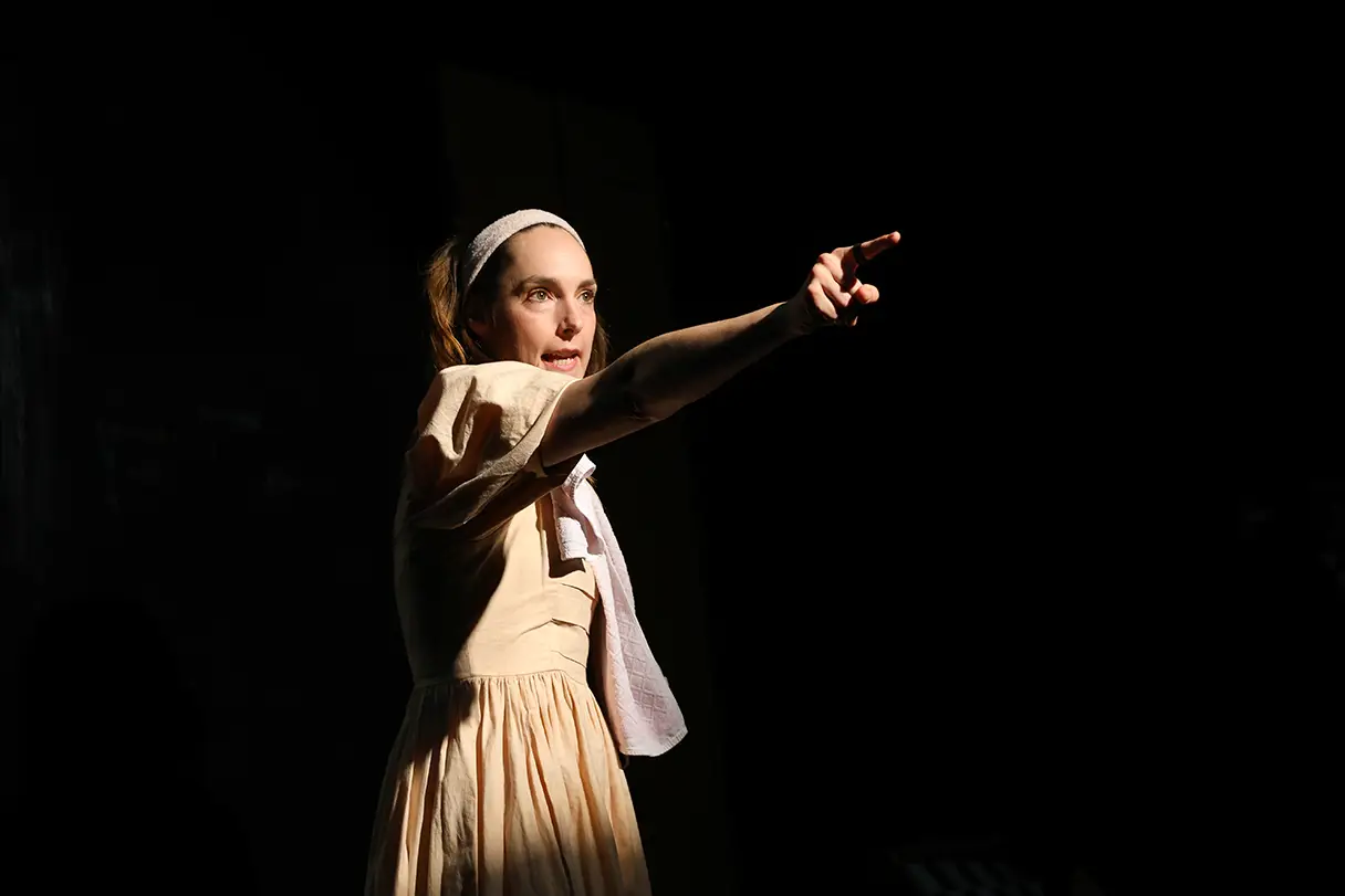 An actress is standing on a stage wearing a yellow dress. The background is completely black. She is pointing forward with her index finger.