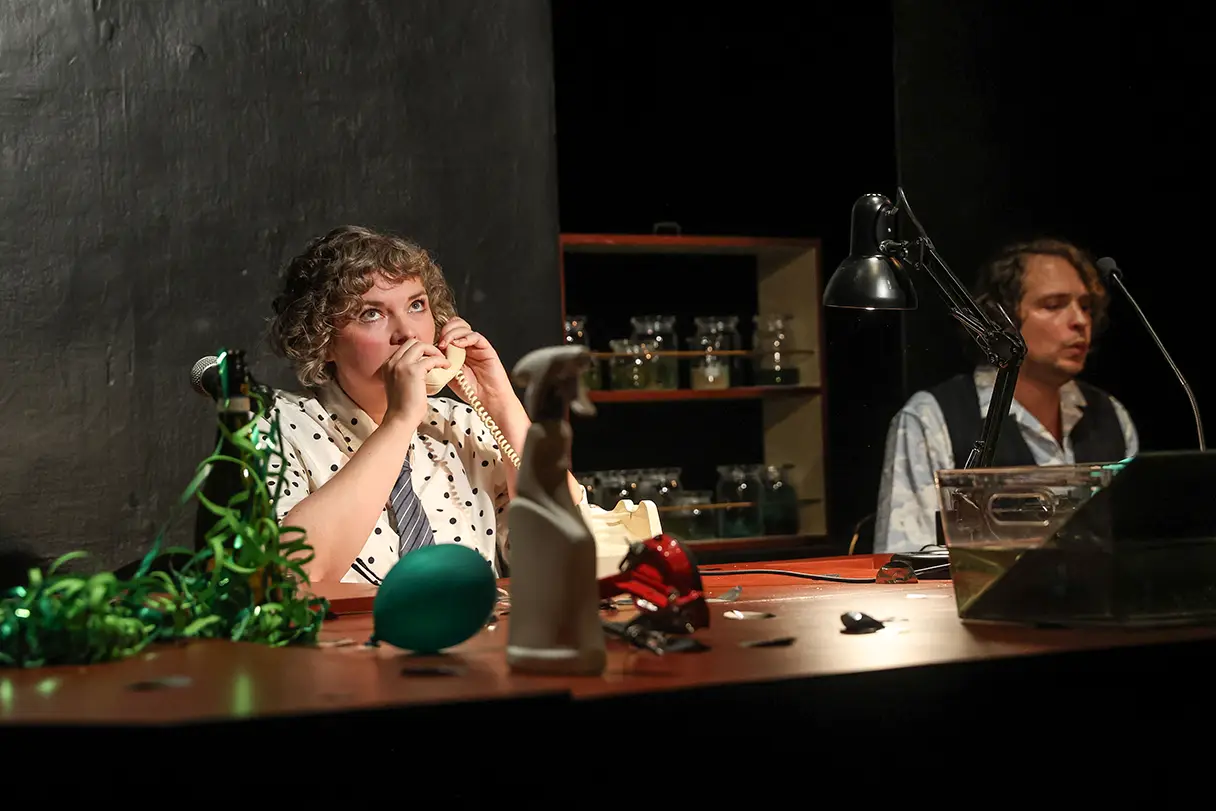 Two actors are sitting at a desk on stage. A woman is on the phone, and a man is sitting next to her. There are various items on the table, including green streamers and a balloon.