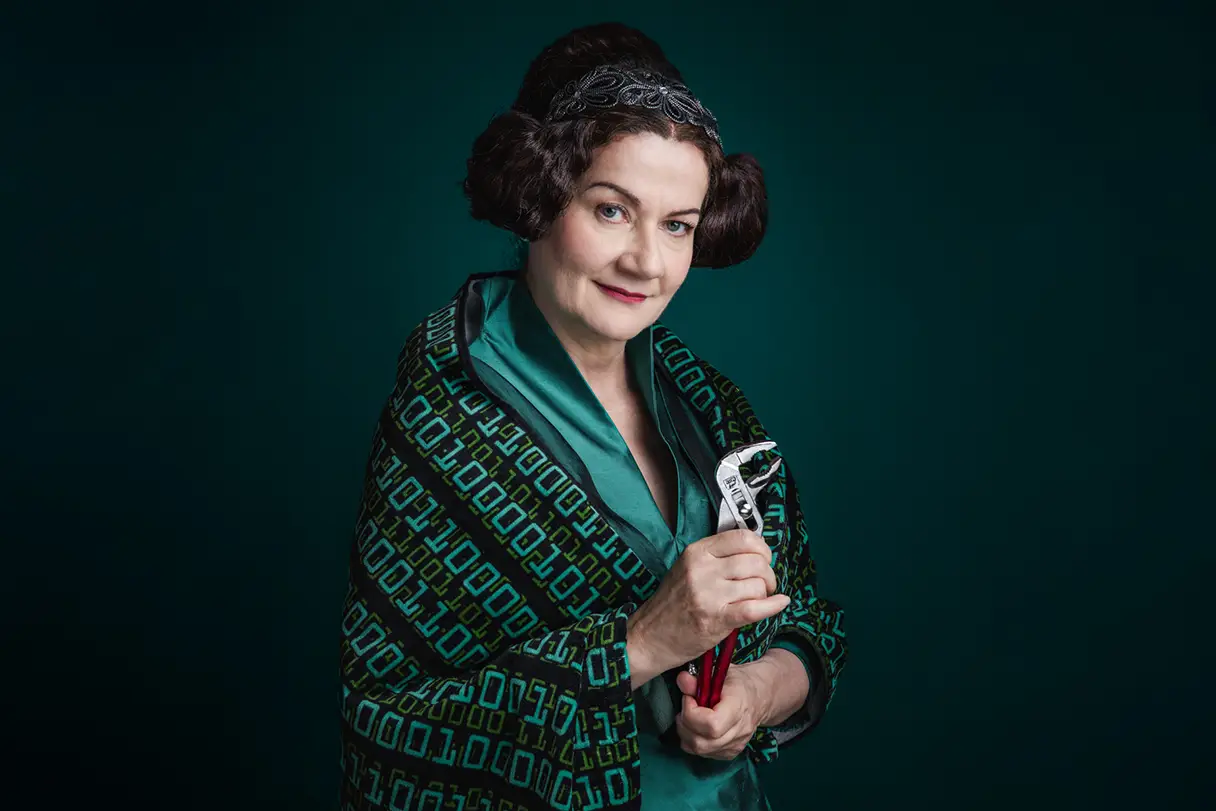 A woman in a green dress with a striking hairstyle is smiling at the camera and holding a pair of pliers.