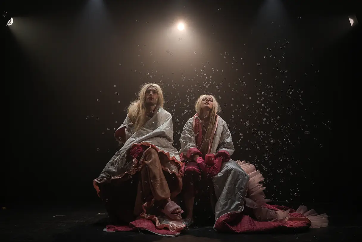 A play at the Theater Drachengasse in Vienna. Two actors sit on a dark stage, wrapped in pink blankets, with countless soap bubbles floating around them.