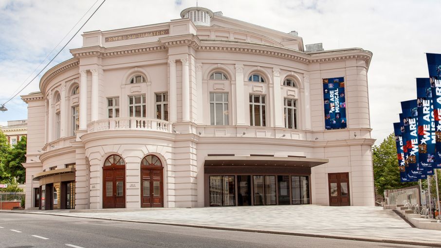 Außenansicht eines historischen Theatergebäudes bei Tageslicht. Die Fassade ist hell und klassizistisch gestaltet, mit hohen Fenstern, Säulen und einem Balkkon. Über dem Eingang ist der Schriftzug 