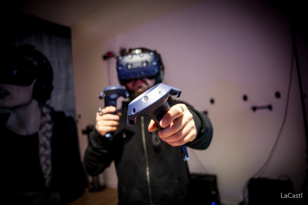 Virtual Escape Room, man holding two game consoles, wearing virtual reality glasses and moving around the room, posters and cables on the wall behind him