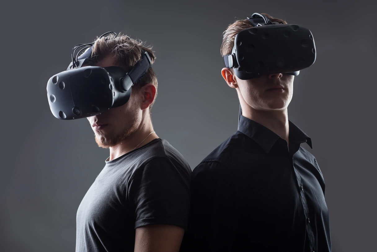 Virtual Escape Room, two men in black T-shirt and shirt standing back to back, both wearing virtual reality glasses, plain gray background