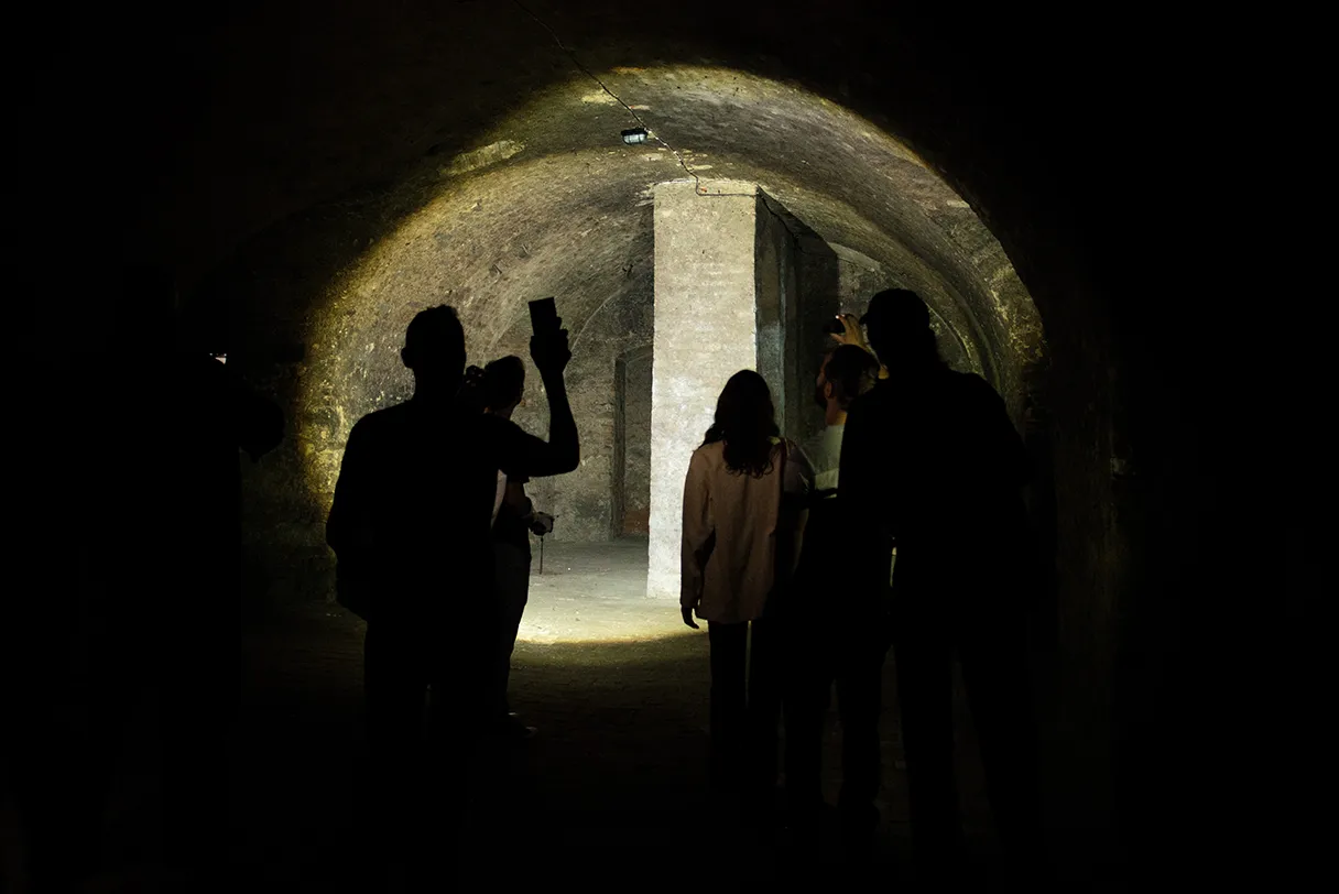 Wien mal anders, Tour in die Wiener Unterwelt, Besucher laufen durch einen dunklen Tunnel, jemand leuchtet mit einer Taschenlampe nach vorn, es ist ein Lichtkegel zu erkennen