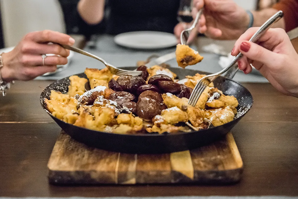 A pan of fresh Kaiserschmarrn is on the table, three people take a piece of Kaiserschmarrn with their forks