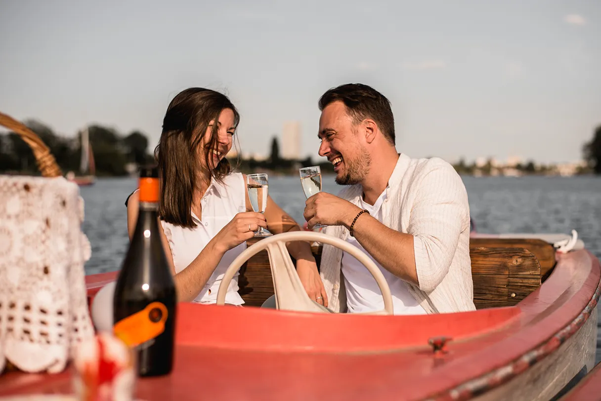 Bootstour mit Wien mal anders. Paar stößt mit Sekt an. Sie lachen. Auf dem Boot steht noch ein Picknickkorb und die Flasche Sekt. Sonniger Tag.