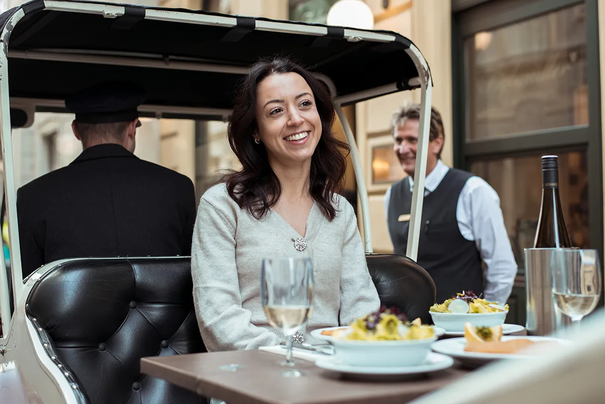 Elektroauto mit Wien mal anders. Eine Frau sitzt in der Kutsche und lacht. Auf dem Tisch vor ihr steht ein Schnitzel von Figlmueller und Pommes.