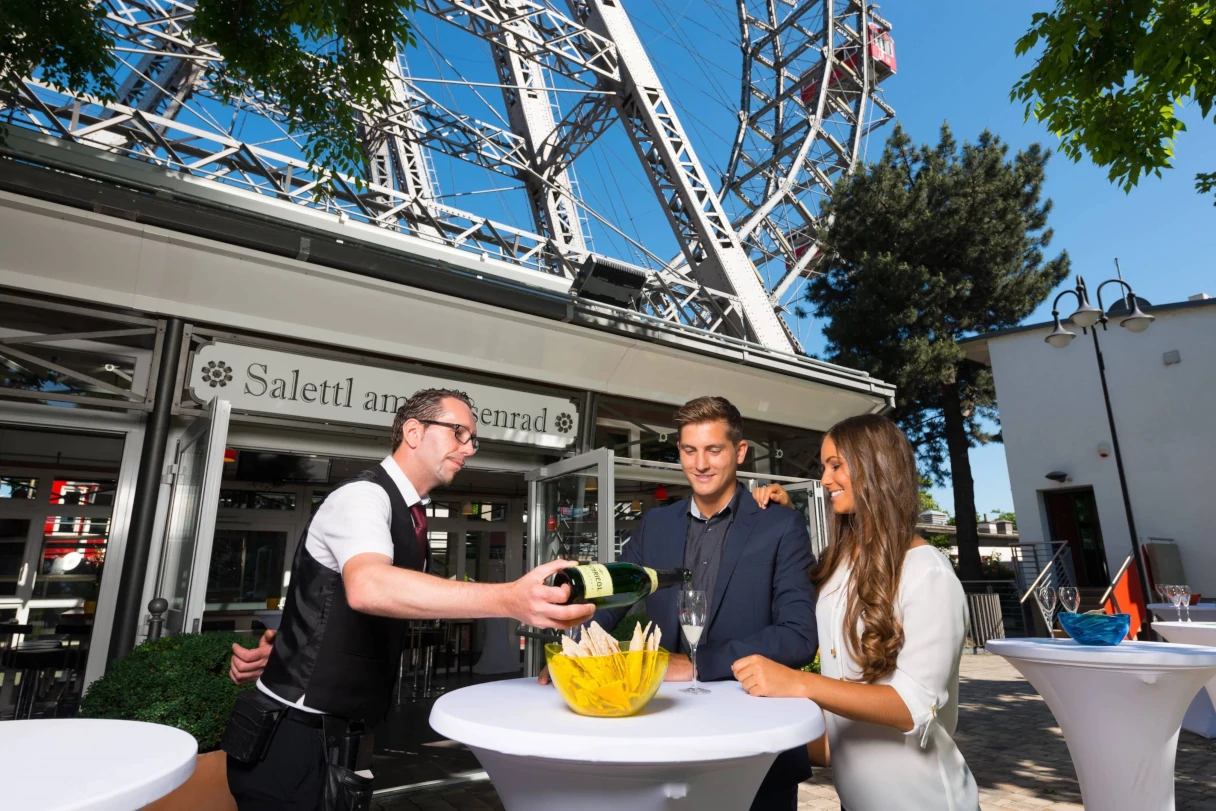 Two guests are being served by a waiter at a white bistro table. They are standing in the Salettl next to the Vienna Giant Ferris Wheel, enjoying a glass of sparkling wine.