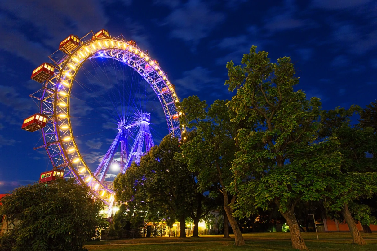 The Vienna Ferris Wheel is lit up at night. There are many trees in the foreground.