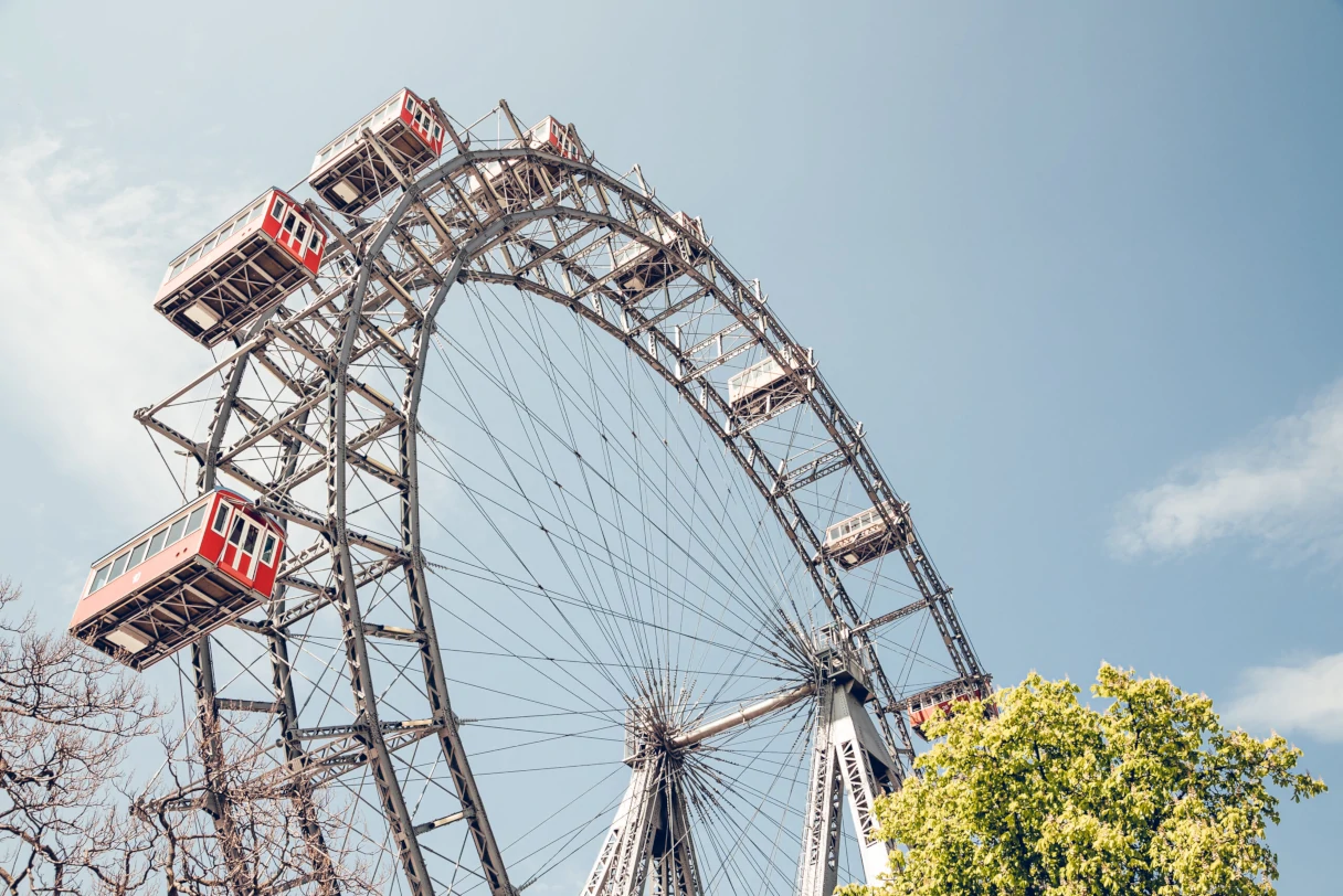 A view of the Vienna Giant Ferris Wheel from the bottom looking up. A tree canopy peeks into the frame at the right edge of the picture. It’s a sunny day with a blue sky and a few wispy clouds drifting by.