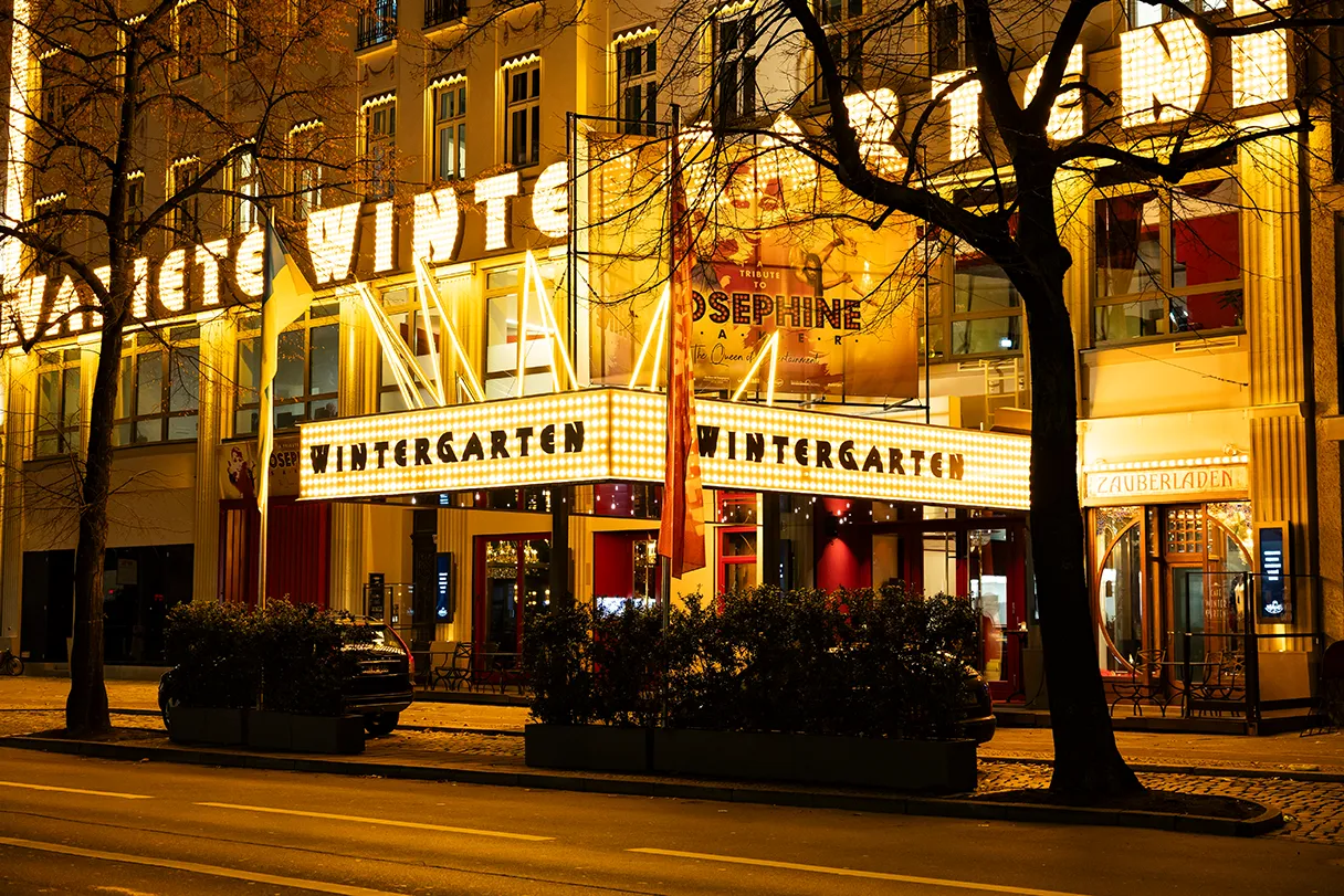 Außenansicht vom Wintergarten Berlin in den Abendstunden. Lichter leuchten golden und an der Häuserwand steht groß Varieté Wintergarten dran.