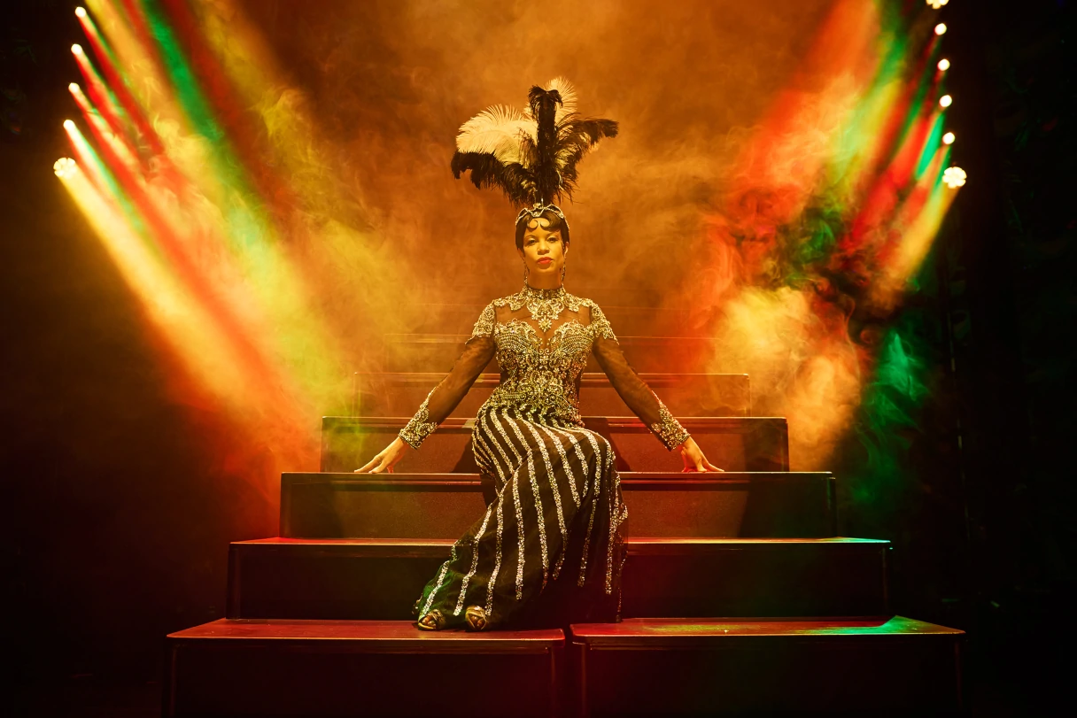 Wintergarten Berlin Varieté, Josephine Baker sits on the show stairs, feather headdress, fog and colorful spotlights on the stage, yellow, green, orange