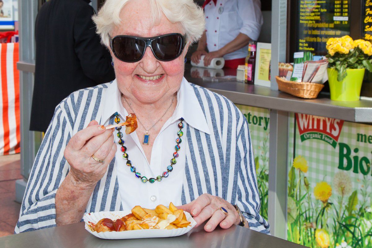 Witty's, Ältere Frau mit schwarzer Sonnenbrille steht am Stehtisch und isst Currywurst mit Pommer, lächelt in die Kamera