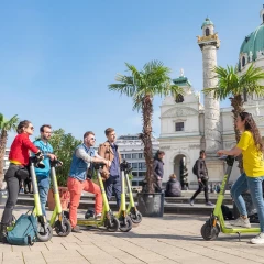 A group of tourists stand on scooters in front of St. Charles Church on Karlsplatz in Vienna. They are sightseeing in a slightly different way. There are palm trees around the small fountain, the sun is shining and the sky is blue.