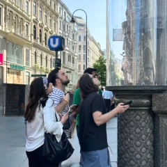 A group of young tourists stand in front of a column on the Graben in Vienna. A subway sign and the shopping street can be seen in the background. The group is trying to solve a riddle.