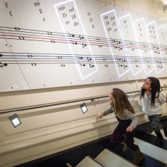 Haus der Musik Vienna, three girls walk up the sound staircase, each step makes a sound, they look to the right at the wall on which a score of notes is attached