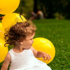 Child in a white dress sits on a green meadow and holds a yellow bunch of balloons in her hand