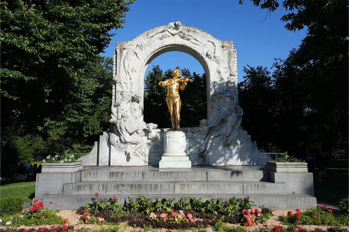 Goldene Statue von Johann Strauss im Wiener Stadtpark. Goldene Statue steht auf einem weißen Sockel und ist umrahmt von Bäumen und Blumen. Sonniger Tag mit blauem Himmel, keine Menschen.