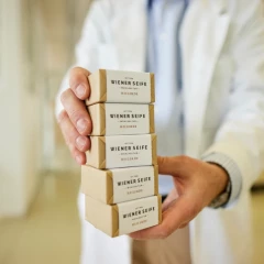 A pile of different Viennese soaps is held in the hands of a man. He is wearing a white coat and has probably just made the soaps.