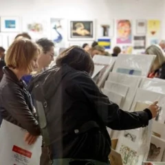 Women stand in a room with many different works of art and look at them. Pictures hang on the walls.