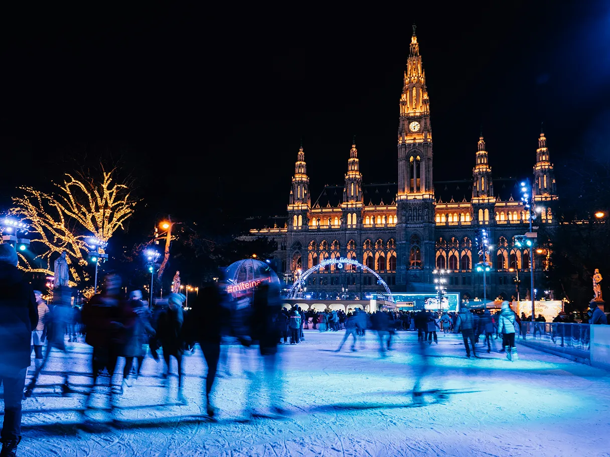 Wiener Eistraum auf dem Wiener Rathausplatz. Menschen fahren Schlittschuh. Das Rathaus ist gelb angeleuchtet und die Eisfläche samt Menschen werden mit blauem Licht angestrahlt.