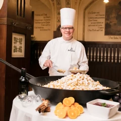 A chef in a white uniform and a large chef's hat stands behind an oversized pan in which Kaiserschmarn is being prepared. He laughs into the camera. Other ingredients and food are placed on the table.ice surface on which a white figure skate is standing. There are some tracks from other skaters on the ice surface.