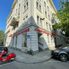 View of the exterior façade and entrance area of the Johann Strauss pharmacy. Beige corner building with red lettering. There are parked cars and a red scooter on the street. Sunny day, blue sky, no people on the street.