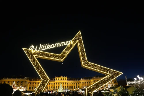 A large golden star at the entrance to the Schönbrunn Christmas Market. The golden Schönbrunn Palace can be seen in the background. A “Welcome” sign hangs from the star.